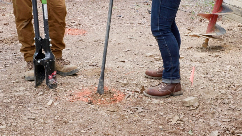 Two people standing near a cirlce marked with orange paint. One person is holding a post hole digger.