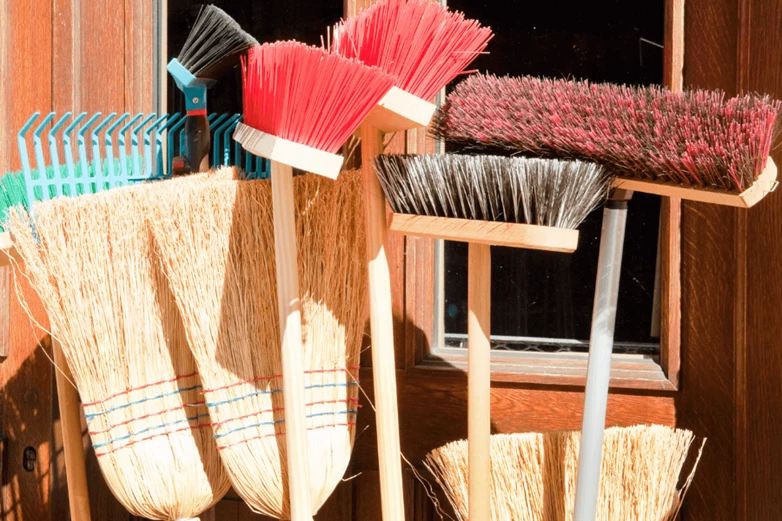 Brushes, straw brooms, push brooms and rakes standing upright agaisnt a wall